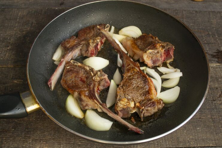 Adding onions to the pan to cook with the lamb chops Adding sliced onions to the skillet with searing lamb chops.