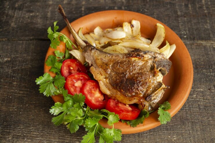 Delicious and juicy bone-in lamb chops ready to serve Plate of cooked bone-in lamb chops with onions, fresh cilantro, and tomatoes.