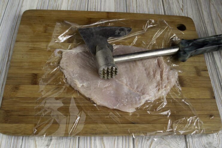A thin piece of chicken fillet being pounded with a meat mallet on a cutting board.