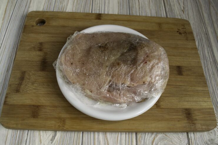 Pounded chicken fillets stacked on a plate, seasoned and covered with plastic wrap for marinating.