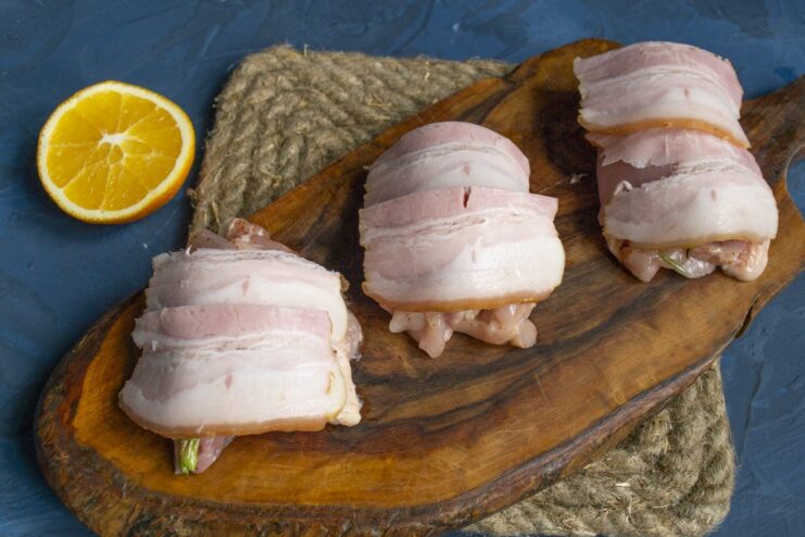 Wrapping the chicken roll with thin slices of bacon for extra flavor and moisture. A chicken roll being carefully wrapped with thin slices of smoked bacon.