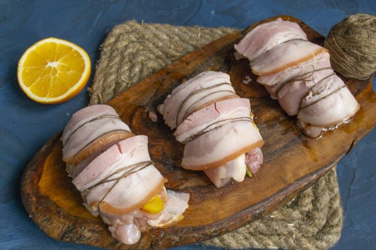 Securing the bacon-wrapped chicken rolls with kitchen twine. Bacon-wrapped chicken rolls being tied with kitchen twine to hold their shape.