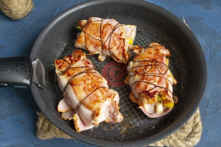 Searing the chicken rolls until golden brown before finishing in the oven. Bacon-wrapped chicken rolls searing in a hot pan, developing a crispy, golden crust.