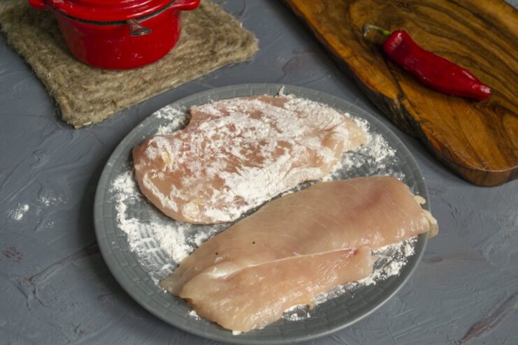 Coating the chicken fillet in flour, the first step of the breading process. A hand dredging a chicken fillet in a plate of white flour.
