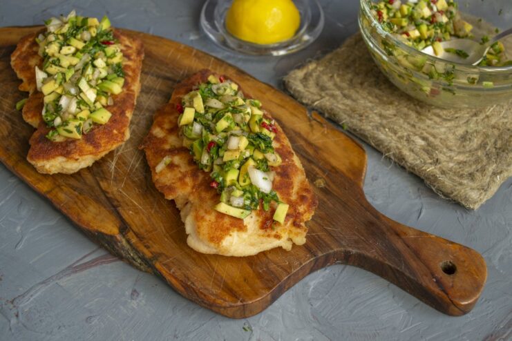 Assembling the dish: topping the crispy schnitzel with fresh avocado salsa. A cooked chicken schnitzel on a wooden board being topped with avocado salsa.