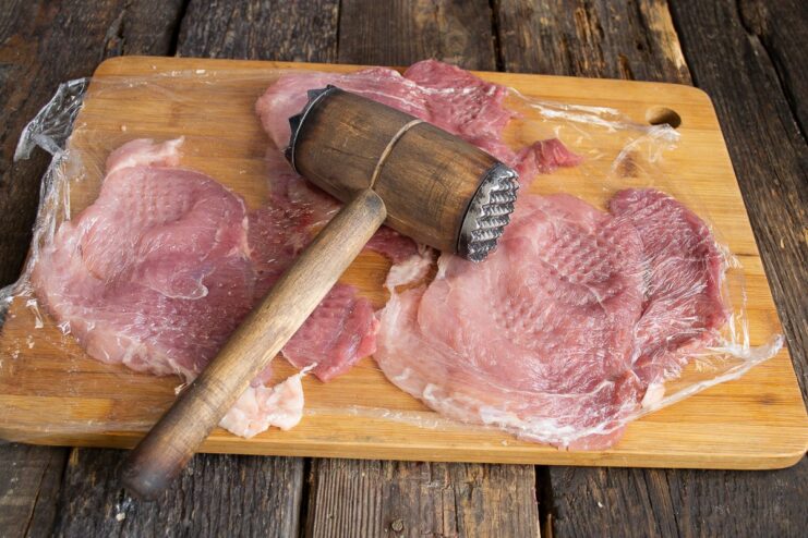 Pounding Pork Cutlets for a Tender Schnitzel Pork cutlets being pounded thin with a meat mallet on a cutting board.