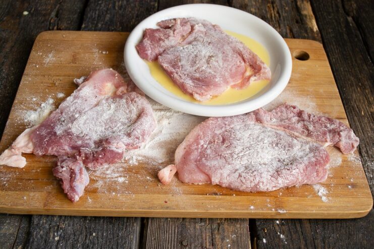 Dipping the Floured Cutlet in Egg Wash A flour-coated pork cutlet being dipped into a bowl of beaten eggs.