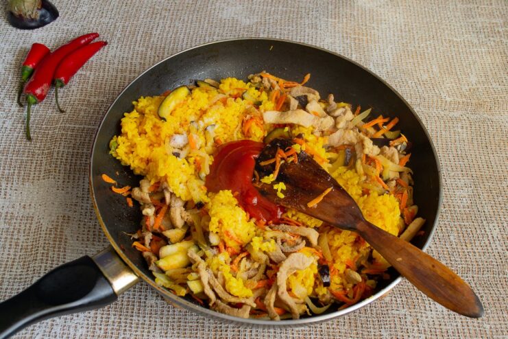 Adding Tomato Paste and Seasoning to Finish the Stew Stirring tomato paste into the pork, eggplant, and rice stew in a pan.