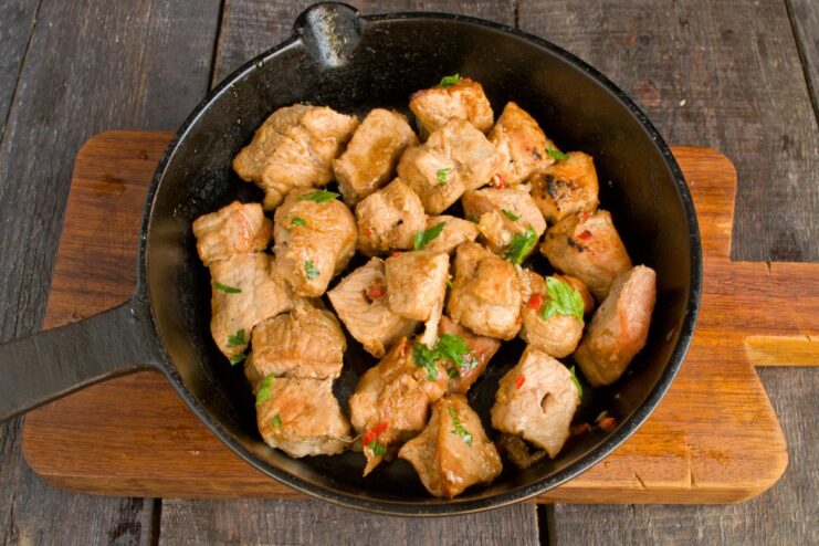 Searing the marinated pork cubes in a hot pan until golden brown. Marinated pork cubes being sautéed in a black skillet.
