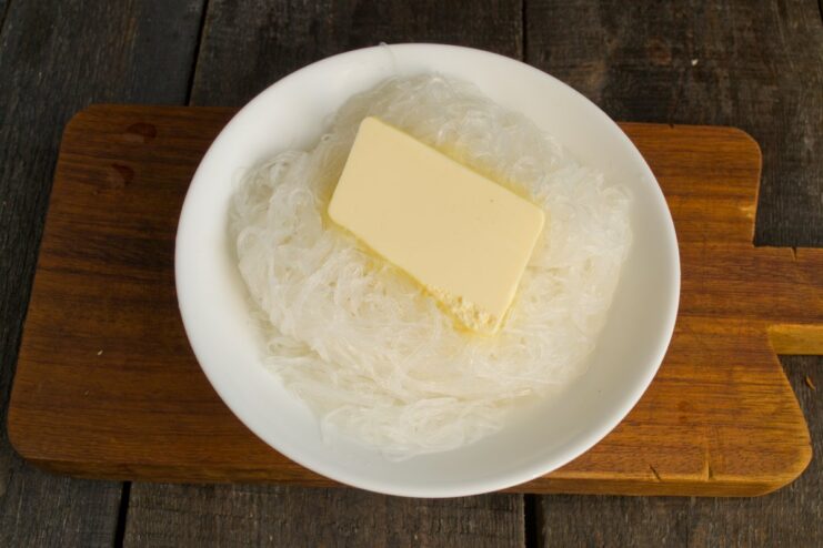 Adding a pat of butter to the hot noodles for extra flavor and creaminess. A pat of butter melting on top of cooked funchoza noodles in a colander.