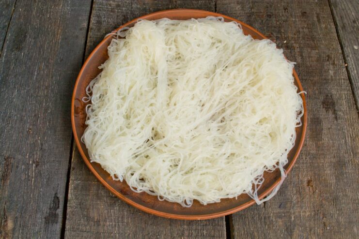 Creating a bed of creamy funchoza noodles on the serving platter. A layer of cooked funchoza noodles spread out on a white serving platter.