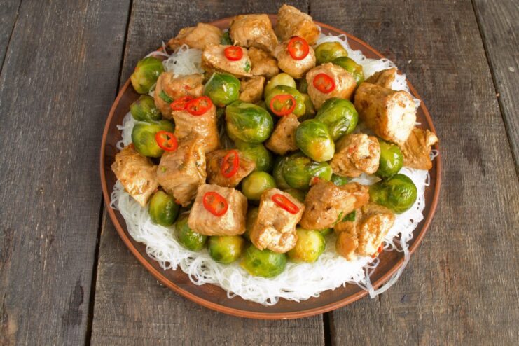 Finishing the dish by adding the savory pork and a sprinkle of spices. Adding the cooked pork and sauce to the platter with noodles and Brussels sprouts.