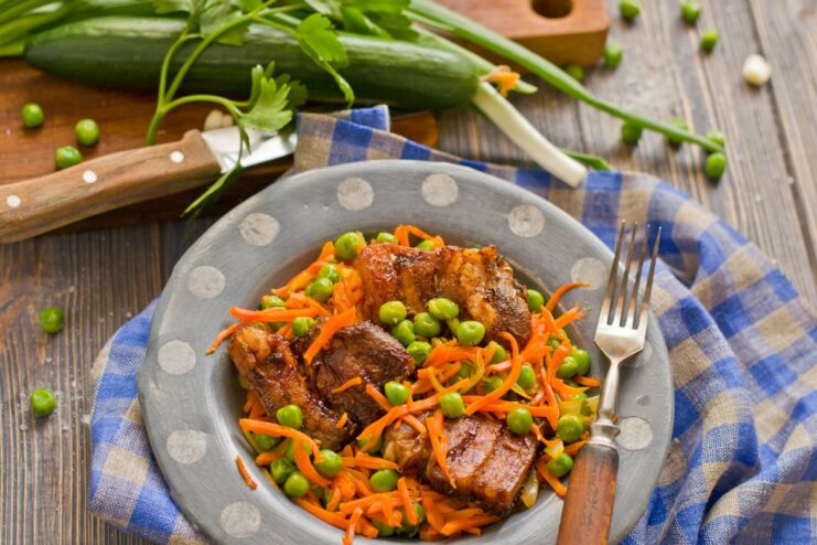 A plate of oven-baked pork with carrots and green peas, garnished with fresh herbs.