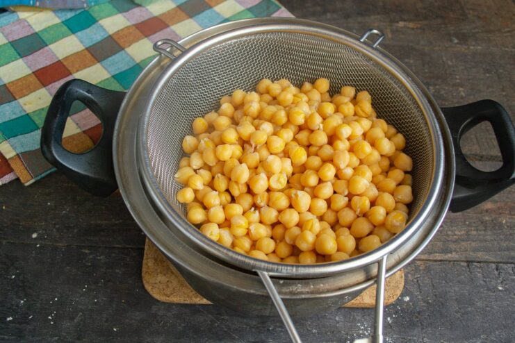 Draining cooked chickpeas is an essential step for the right curry texture. Cooked chickpeas being drained in a metal sieve over a sink.