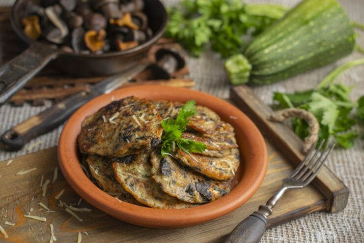 A plate of golden-brown zucchini fritters with parmesan and mushrooms, garnished with fresh herbs.
