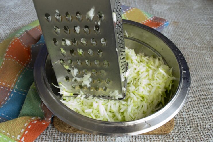 Step 2: Grating the peeled zucchini to prepare it for the fritter batter. Shredded zucchini in a white bowl after being grated on a box grater.