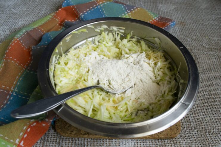 Adding flour to the mixture to create the perfect fritter batter consistency. Spoonful of white flour being added to the zucchini fritter batter in a bowl.