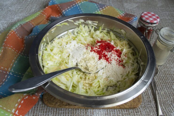 Flavoring the batter with spices like paprika and fenugreek for a rich taste. Spices being added from a spoon into the zucchini fritter batter.