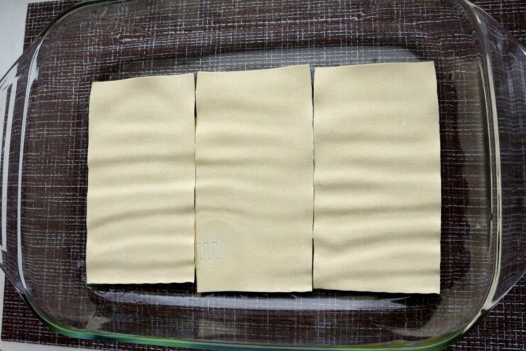 Assembling the lasagna: Starting with the first layer of pasta Dry lasagna sheets being placed in a single layer in a greased glass baking dish.