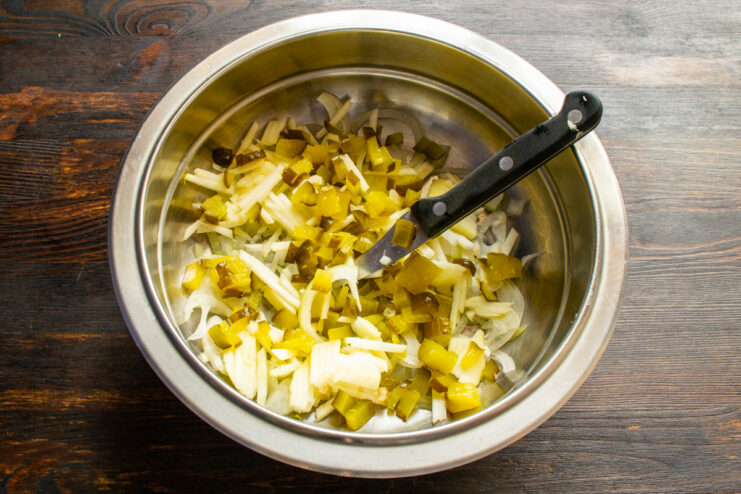 Dicing fermented pickles for a tangy crunch in the salad. A hand dicing salted pickles into small cubes on a cutting board.