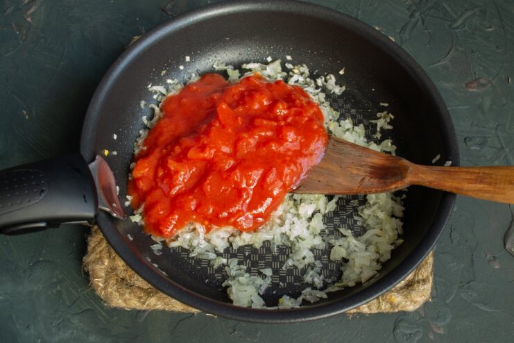 Adding Canned Tomatoes and Sugar to the Sautéed Onions Canned tomatoes and sugar being added to sautéed onions in a pan.
