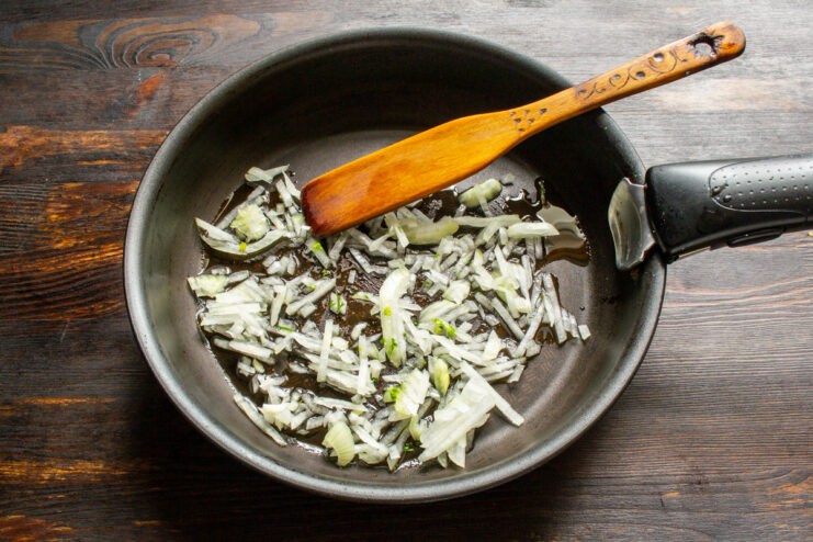 Sautéing finely chopped onions in a pan as the first step of the recipe. Finely chopped onions being sautéed in a large pan with oil.