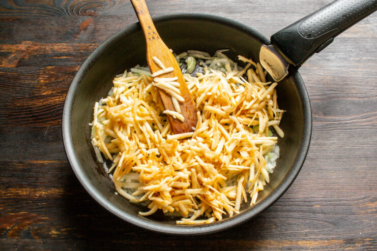 Adding grated apples to the sautéed onions in the pan. Grated apples being added to a pan with sautéing onions.