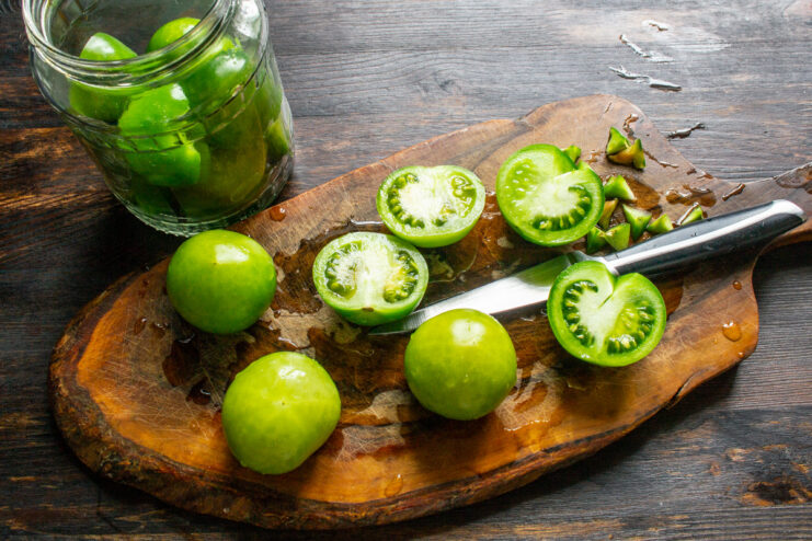 How to Prep Green Tomatoes for Pickling - Halving and Coring A hand holding a knife, cutting a green tomato in half on a wooden cutting board.