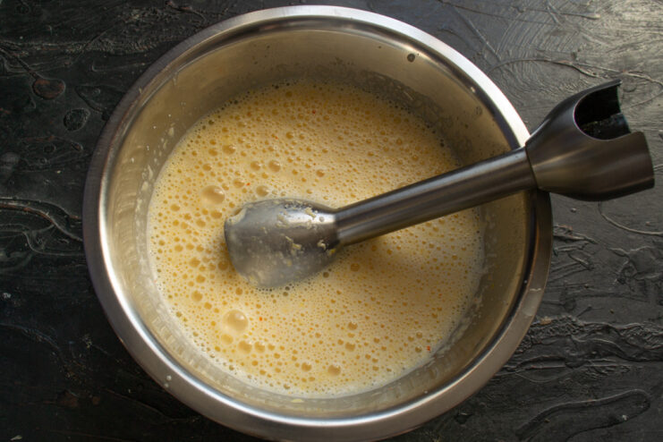 Using an immersion blender to create a smooth, homogeneous batter base. An immersion blender pureeing the orange, whey, eggs, and oil in a bowl.