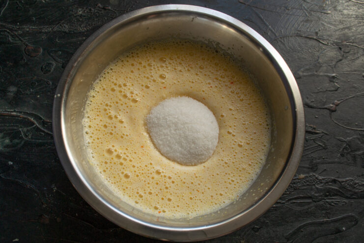Adding sugar to the blended wet ingredients for the orange mannik. Granulated sugar being poured into the blended orange mixture.
