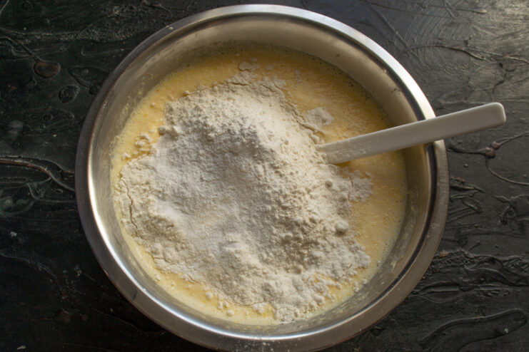Thoroughly mixing the mannik batter to ensure a smooth consistency. A whisk mixing the semolina cake batter in a bowl until smooth.
