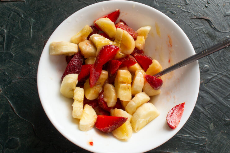 Sliced strawberries and bananas being gently tossed in a honey and lemon sauce in a bowl.