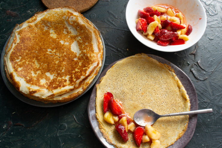 A lacy crepe being rolled up with fruit salad filling, with sauce drizzled over the top.