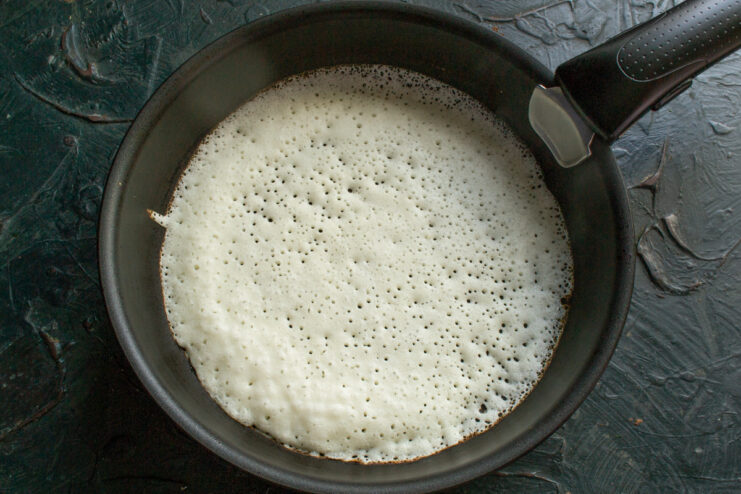 A thin, lacy crepe cooking in a hot skillet, with bubbles forming on the surface.