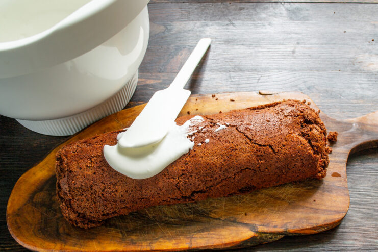 Frosting the roll with a light and tangy smetana cream. A spatula spreading white smetana cream over the top of the chocolate sponge roll.