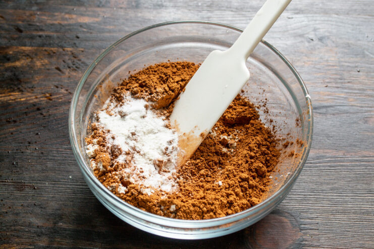 Combining the dry ingredients for the chocolate sponge cake. A bowl containing cocoa powder, oat flour, and baking powder being mixed with a whisk.