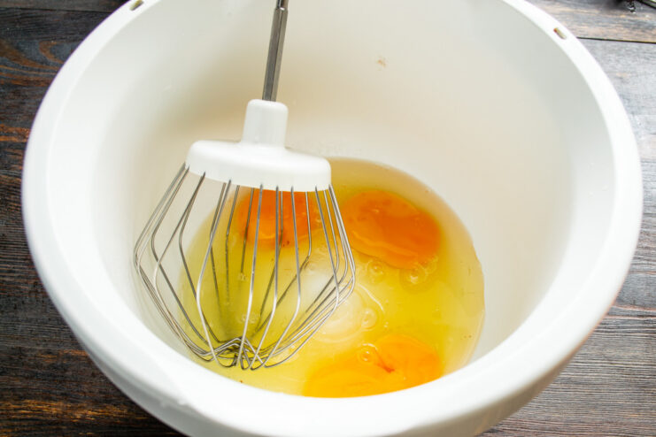 Starting the sponge cake batter by cracking eggs into a mixer bowl. Three eggs cracked into the glass bowl of a stand mixer.
