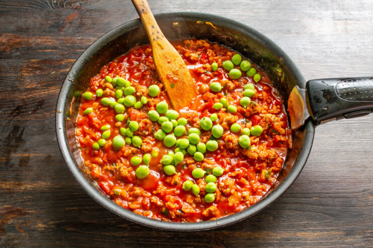Adding a Pop of Color and Sweetness with Green Peas Green peas being added to the simmering meat sauce in the pan.