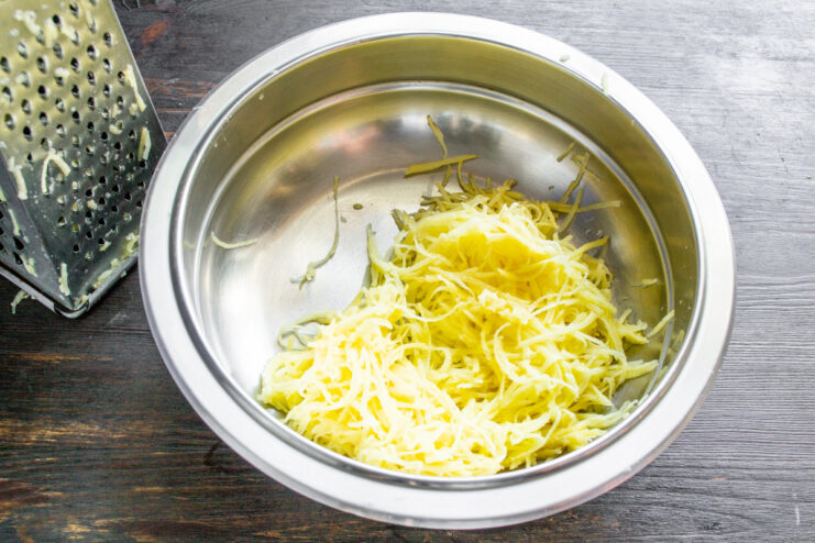 Peeled raw potatoes being grated on a box grater into a bowl.