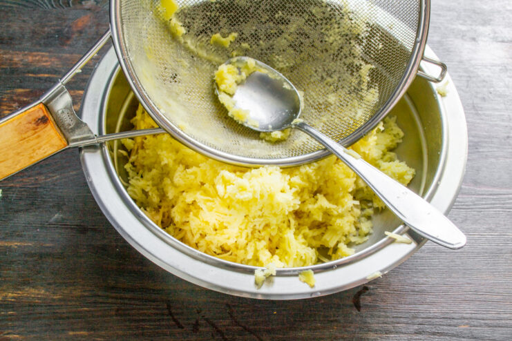 A hand using a spoon to press cooked potato cubes through a sieve.