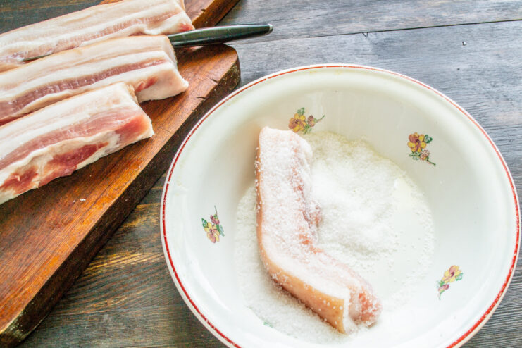 Starting the curing process: Generously coating the pork belly with salt. Pieces of pork belly being coated with coarse sea salt.