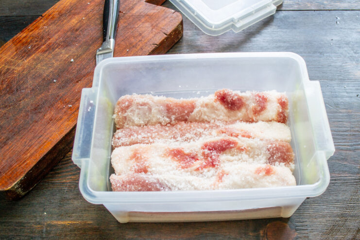 Packing for curing: Ensuring the pork belly is fully covered in salt. Salt-coated pork belly pieces packed tightly into a plastic container.