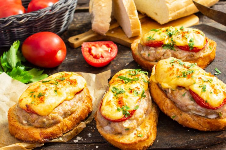 A plate of baked open-faced sandwiches with minced meat, melted cheese, and tomato, garnished with fresh parsley.