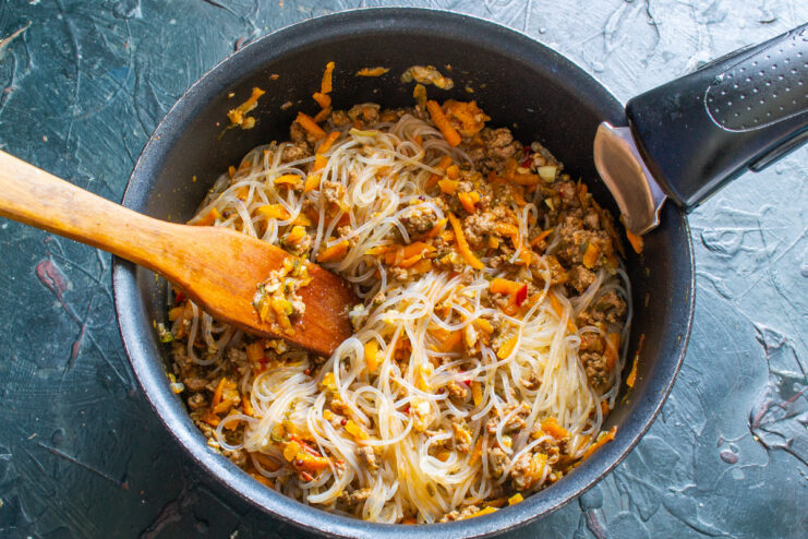 Mixing the Noodles and Sauce - Ensure the noodles are well-coated. Funchoza noodles being thoroughly mixed with the meat and vegetable sauce in a pan.