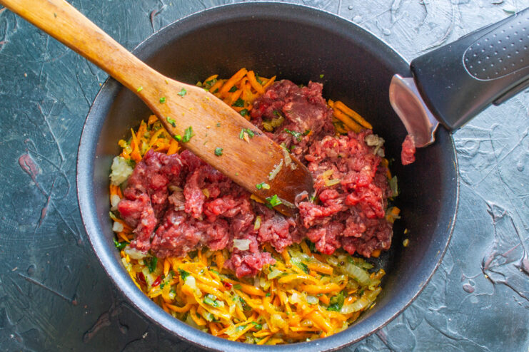 Browning the Ground Beef - Break it up with a spatula as it cooks. Ground beef being added to the sauté pan with vegetables and stirred.