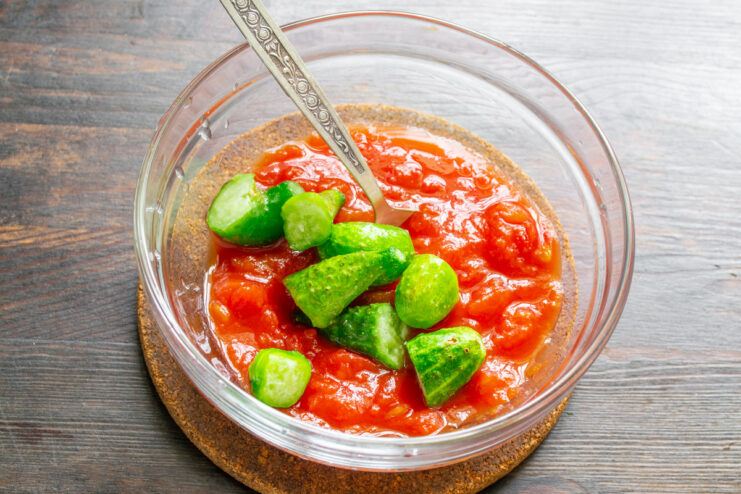 Adding Freshness: Cucumbers and Garlic for Gazpacho Finely chopped cucumbers and minced garlic being added to a bowl of canned tomatoes.