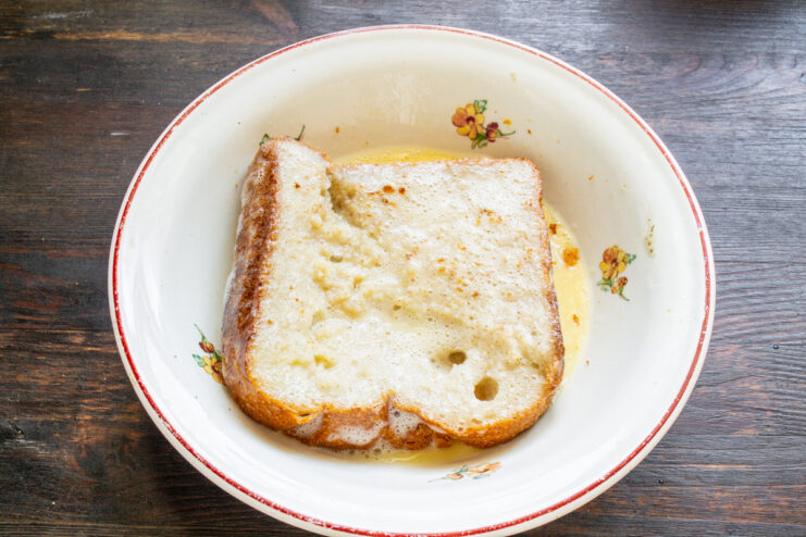 Soaking Bread Slices in Egg Mixture for Even Cooking A slice of bread being dipped into a bowl with a yellow egg and milk mixture.