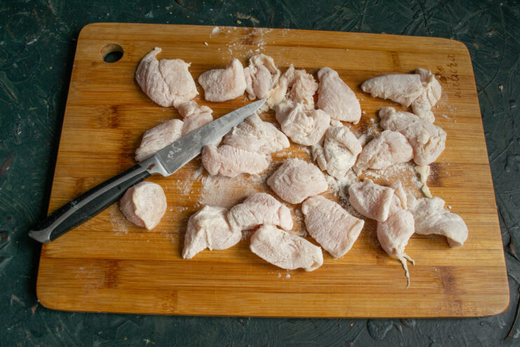 Cutting chicken breast into large chunks for a hearty soup. Raw chicken breast fillet being cut into large chunks on a cutting board.