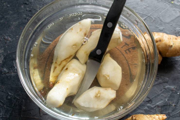 Preparing Jerusalem Artichokes for Draniki Hands peeling a Jerusalem artichoke with a vegetable peeler over a bowl of water.