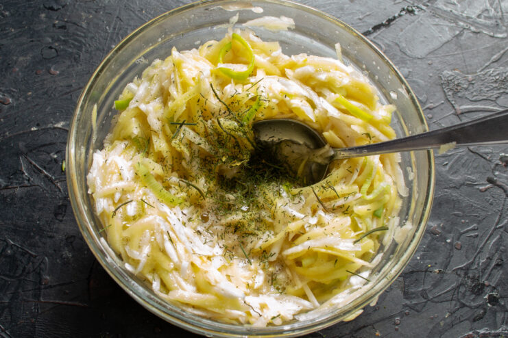 Seasoning the Batter with Dried Dill Adding a spoonful of dried dill to the draniki batter in a bowl.